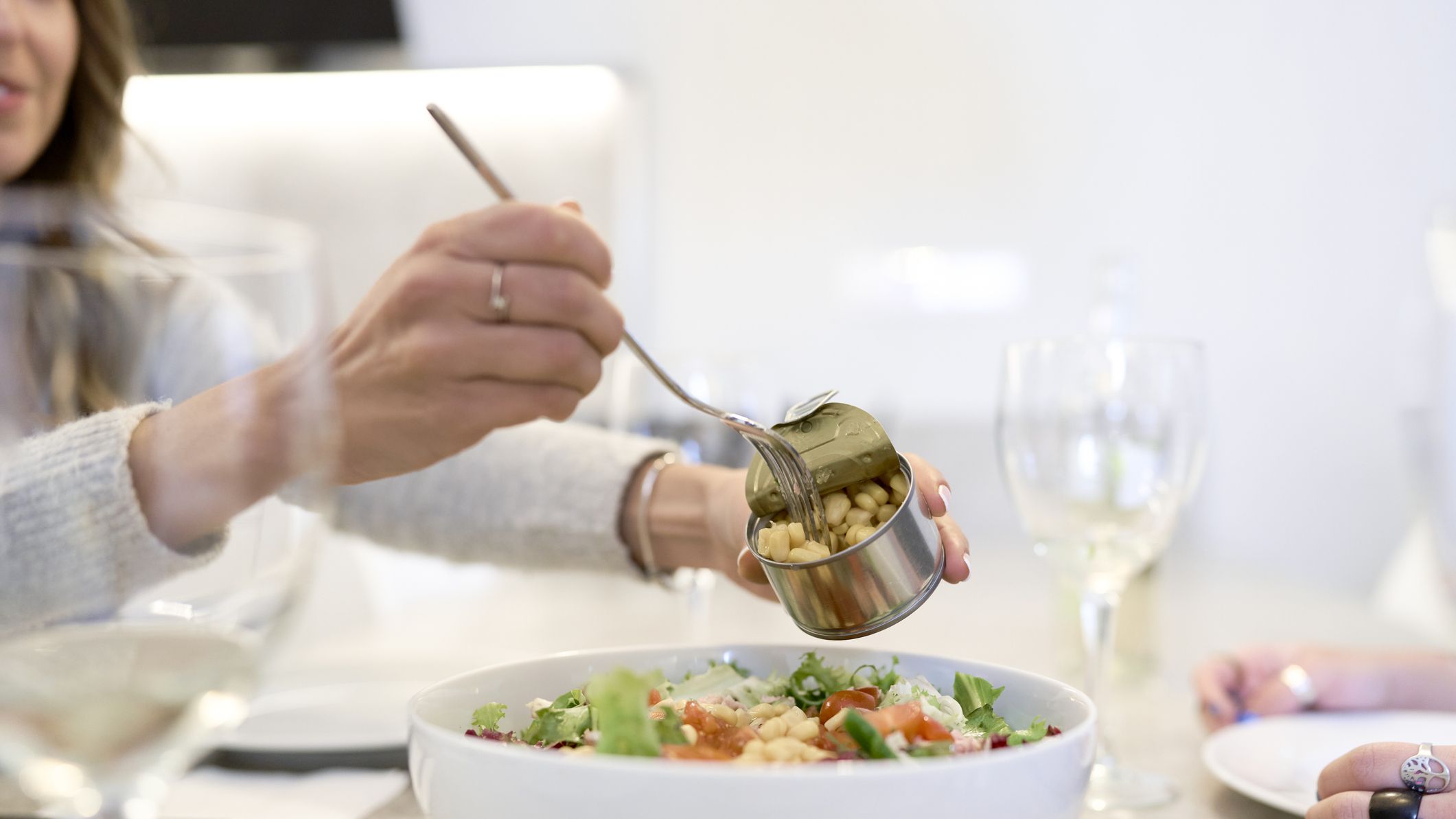 Woman at a set dining table uses a fork to decant white beans from a tin into a salad bowl