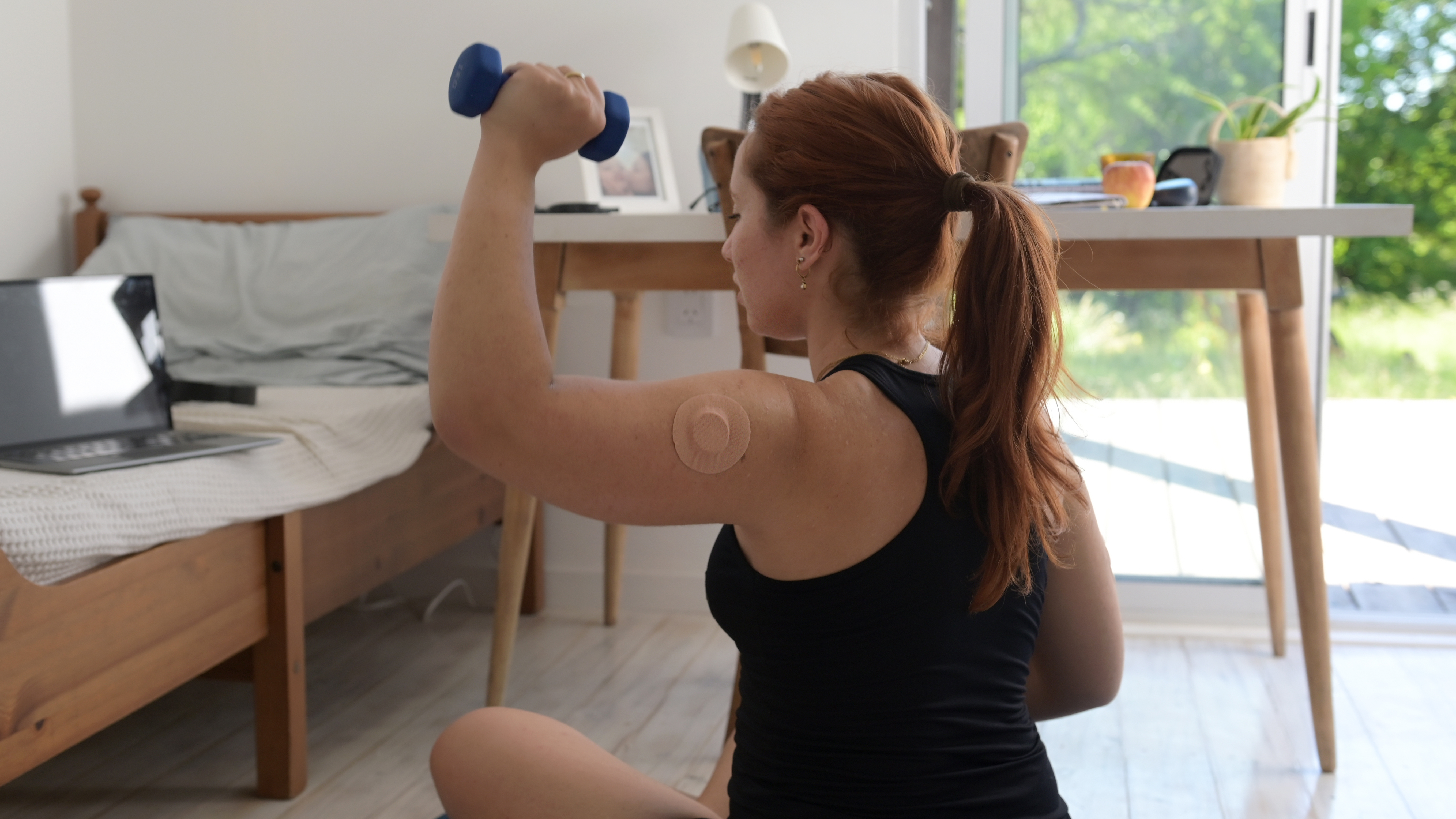 Una donna con un monitor del glucosio al braccio tiene un manubrio leggero in mano. Siede, con il manubrio sollevato all'altezza della testa.